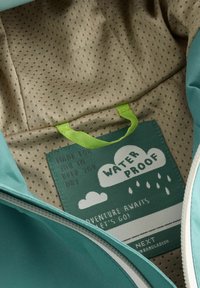 Étiquette de vêtement à l'intérieur d'une veste verte avec texte "Waterproof," nuages, gouttes de pluie, "Adventure awaits, let's go," et "Made for you to keep you dry."