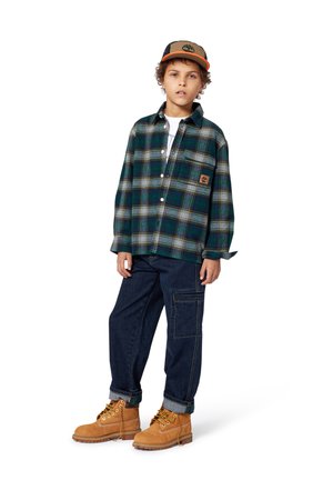 Chemise en flanelle à carreaux verts et bleus, avec boutons, poche et patch logo ; pantalon en denim bleu foncé ; bottes beiges avec lacets jaunes et revers roulés.