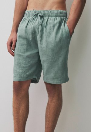 Mann trägt hellgrüne Shorts aus strukturiertem Stoff mit elastischem Bund und Kordelzug, steht mit den Händen in den Taschen vor einem einfarbigen Hintergrund.