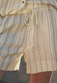 Lys beige shorts lavet af tekstureret stof med vertikale sorte striber. Har en justerbar snøre i taljen med dekorative perler.