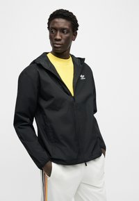 Άνδρας που φοράει μαύρο μπουφάν Adidas με φερμουάρ πάνω από κίτρινο πουκάμισο, λευκό παντελόνι με πλευρικές ρίγες, στέκεται με τα χέρια στις τσέπες κοιτάζοντας προς τα πλάγια.