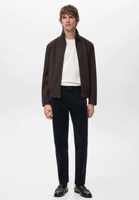 Chaqueta marrón con cremallera sobre una camisa blanca, combinada con pantalones oscuros y zapatos negros. Diseño sencillo y moderno con un ajuste estructurado.