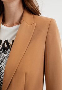 Brown tailored blazer with sharp lapels, smooth fabric texture, and a relaxed fit, layered over a graphic white t-shirt.