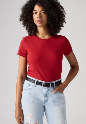 Жена с къдрави коси, облечена в червена тениска Levi's, светли дънки с разкъсвания и черен колан, стои с една ръка в джоба си.