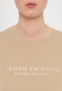 Béžové bavlněné tričko s kulatým výstřihem a zlatým fóliovým potiskem "ARMANI EXCHANGE MILANO / NEW YORK" na hrudi.