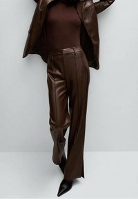 Osoba nosząca ciemnobrązowe skórzane dopasowane spodnie, pasującą skórzaną kurtkę i spiczaste czarne botki, stoi na tle jasnoszarym.