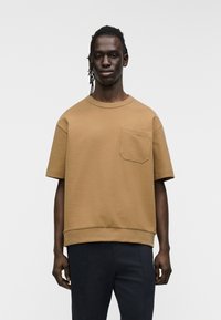 Καφέ βαμβακερό κοντομάνικο μπλουζάκι με στρογγυλή λαιμόκοψη και τσέπη στο στήθος, με ίσια γραμμή στο κάτω μέρος και άνετη εφαρμογή.