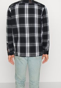 Svartvit rutigt skjorta med långa ärmar, knappmanschetter och rak fåll, i kombination med ljusblå jeans. Strukturerad tyggdetalj synlig.