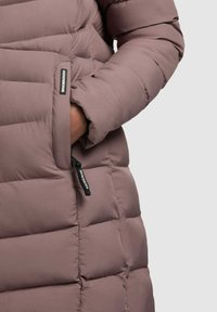 Gros plan de la main gauche dans la poche d'une veste matelassée mauve avec zip noir et petite étiquette de marque.