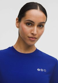 Sötét hajú nő, akinek a haja hátrafogva van, élénk kék inget visel, amelynek gallérja közelében három kis fehér logó található.