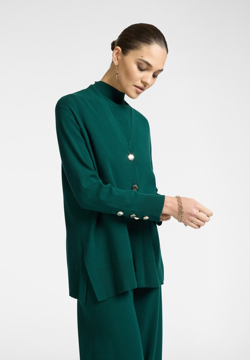 Vihreä neulepaita, jossa on pitkät hihat, korkea kaulus ja napit hihansuissa; yhdistetty samanvärisiin housuihin.