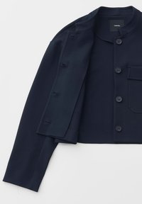 Giacca corta blu scuro con collo tondo, bottoni frontali, tasca sul petto con pattina e maniche lunghe posata distesa su sfondo bianco.