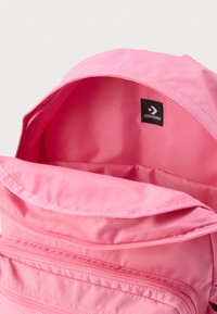 Zaino in tessuto rosa con un compartimento principale con cerniera e un'etichetta con logo interna. Presenta una texture liscia e un design semplice.