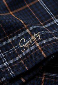 Granatowa tkanina w kratkę z pomarańczowymi i jasnoniebieskimi liniami, zdobiona złotym haftowanym logo "Superdry". Miękka tekstura, widoczny splot.