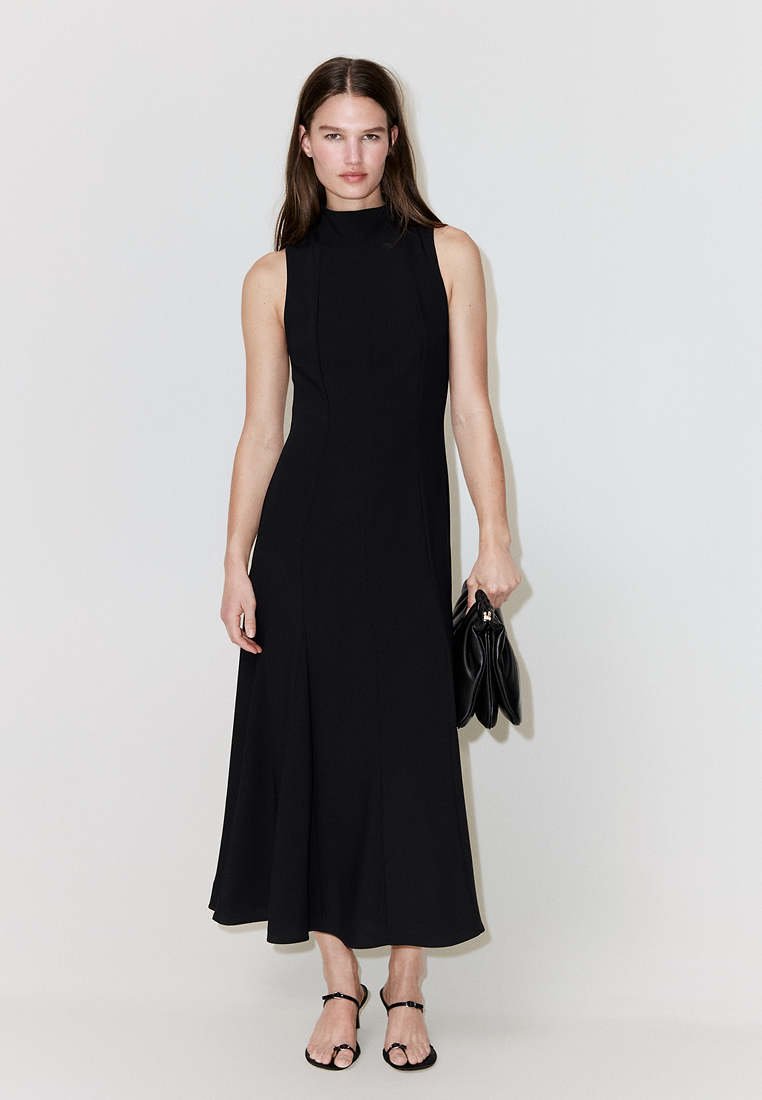 Jeune femme portant une robe midi noire sans manches et des sandales noires, tenant une pochette noire pliée devant un fond clair uni.