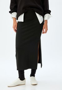 Zwarte, getailleerde midi-rok met een zijsplit. Gecombineerd met een wit shirt en een zwarte trui. Witte sneakers maken de look compleet.