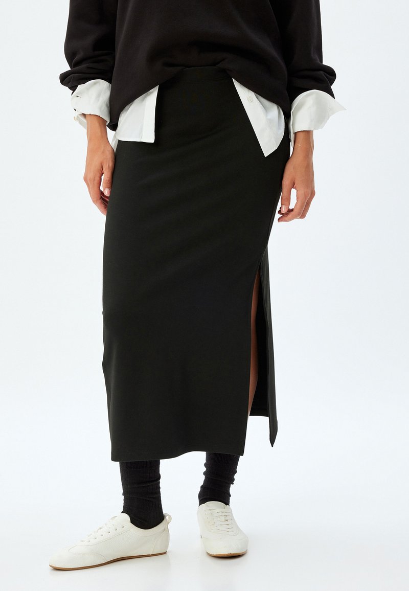 Jupe midi ajustée noire avec une fente sur le côté. Associée à une chemise blanche et un sweater noir. Des baskets blanches complètent le look.