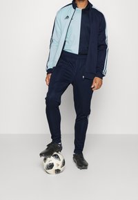 Blå och mörk marinblå idrottsoverall med vita ränder, dragkedjefickor och svarta fotbollsskor. Personen står med en fotboll vid sina fötter.
