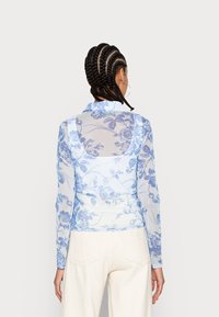Blusa transparente de mangas longas em azul claro com padrões florais. Apresenta um colarinho e um design ajustado, combinada com calças de cor creme.