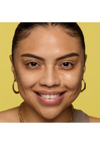 Personne souriante à la peau lisse, portant des boucles d'oreilles créoles en or et un collier en or, sur un fond jaune uni.