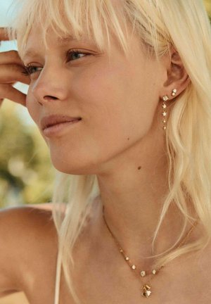 Mlajša ženska s svetlo blond lasmi, ki nosi zlate cvetlične uhane in ujemajoč se nakit, mirno gleda v stran na prostem v mehki naravni svetlobi.