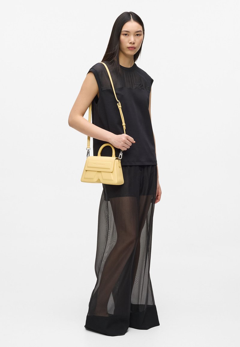 Haut noir sans manches avec un voile transparent, associé à un pantalon noir transparent. Porte un petit sac à main en cuir jaune au design structuré.