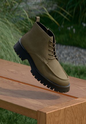 Olivengrønne suedestøvler til ankelen med sorte robuste såler, med snøredesign og trekkhank på hælen, vist over en treoverflate.