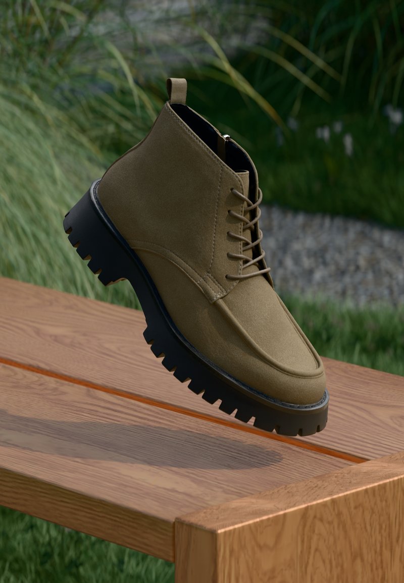 Alyvuogių žalios zomšos puskojinės su juodais tvirtais padais, su raištelių uždarymo dizainu ir traukimo kilpa ant kulno, padėtos virš medinės paviršiaus.