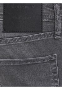 Jeans de mezclilla gris con un parche de cuero negro con texto en relieve. Incluye detalles de costura y un bolsillo trasero. Textura suave.