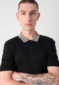 Fekete póló, textúrált világosszürke gallérral, három gombos felső részével és rövid ujjaival. Karján bonyolult tetoválásokkal.