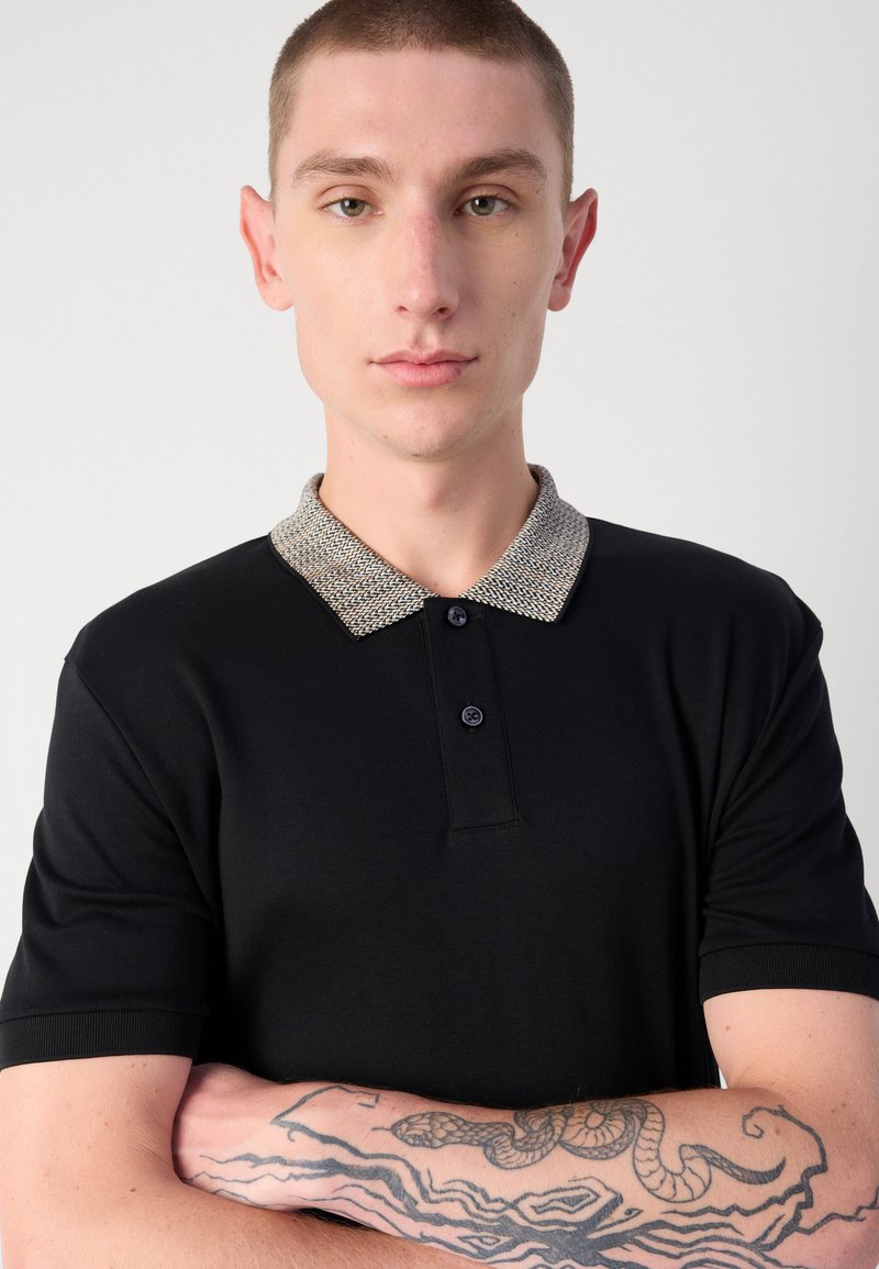 Schwarzes Poloshirt mit einem strukturierten hellgrauen Kragen, ausgestattet mit einer Knopfleiste mit drei Knöpfen und kurzen Ärmeln. Arm mit kunstvollen Tattoos bedeckt.