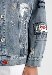 Džínová bunda s stříbrnými flitrovými písmeny "YF", vyšitým nášivkou s červenými rty, černým vyšíváním a opotřebovanými detaily na rukávu.