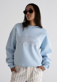 Sudadera azul claro con cuello redondo, corte oversized y logo bordado de "Prohibido". Combinada con pantalones de rayas beiges.