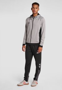 Chaqueta gris con cremallera y capucha estampada, con detalles en negro, combinada con pantalones negros que tienen paneles laterales grises y detalles en chevron blanco.
