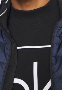 Sudadera negra con texto blanco debajo de una chaqueta de cremallera color marino. La chaqueta presenta una textura suave y un borde blanco en contraste alrededor del cuello.