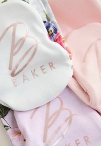 Minkštos audinio prekės pastelinių spalvų su gėlių raštais ir metaliniu rožiniu „B Baker“ logotipu ant jų.