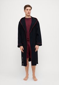 Svart badekåpe i frotté med røde detaljer, lange ermer og hette, brukt over en bordeaux t-skjorte med korte ermer og shorts. Bare føtter.