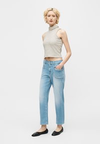 Νεαρό άτομο με κοντά σγουρά ξανθά μαλλιά, που φοράει αμάνικο ριγέ κοντό τοπ, ανοιχτό μπλε τζιν και μαύρες μπαλαρίνες, στέκεται με το ένα χέρι στην τσέπη.