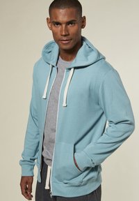Hellblauer Zip-Hoodie aus weichem Material mit einer Kapuze mit Kordelzug, versehen mit Fronttaschen und gerippten Bündchen. Kombiniert mit einem grauen T-Shirt.
