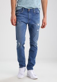 Blå denimjeans med slitna detaljer, slim fit-design, femficksstil och en urtvättad look. Matchas med vita sneakers.
