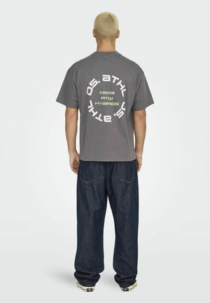 Gray cotton t-shirt with oversized fit, featuring circular graphic text in white and neon green on the back. Worn with dark denim pants.