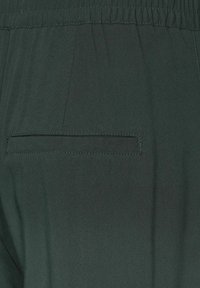 Mörkgrön tyg med en jämn textur, med en elastisk midja och en bakficka med sömnadsdetaljer.