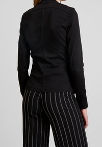 Giacca nera aderente con colletto alto, maniche lunghe e cuciture a vista. Abbinata a pantaloni a gamba larga a righe bianche e nere.