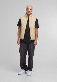 Beige quilted vest with high collar over a black t-shirt, paired with dark denim pants and white sneakers. Plain design, no visible patterns.