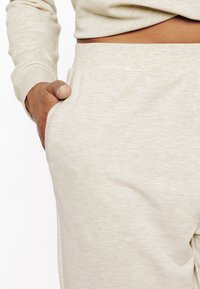 Tissu de sweat-shirt beige clair avec une texture douce. Dispose d'une ceinture ajustée et d'une poche latérale, partiellement recouverte par une main.