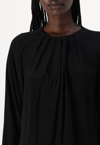 Blusa nera con un design drappeggiato, caratterizzata da pieghe lungo il colletto. Realizzata in tessuto morbido, con maniche lunghe e colletto arrotondato.