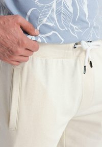 Pantaloni della tuta beige con coulisse, vita a coste e tasche laterali, presentati con una camicia blu a motivo floreale. Texture liscia.
