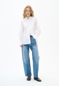 Weißes Hemd mit Piquet-Details, langen Ärmeln und Stehkragen, kombiniert mit lockeren hellblauen Jeans und schwarzen Ankle-Boots.