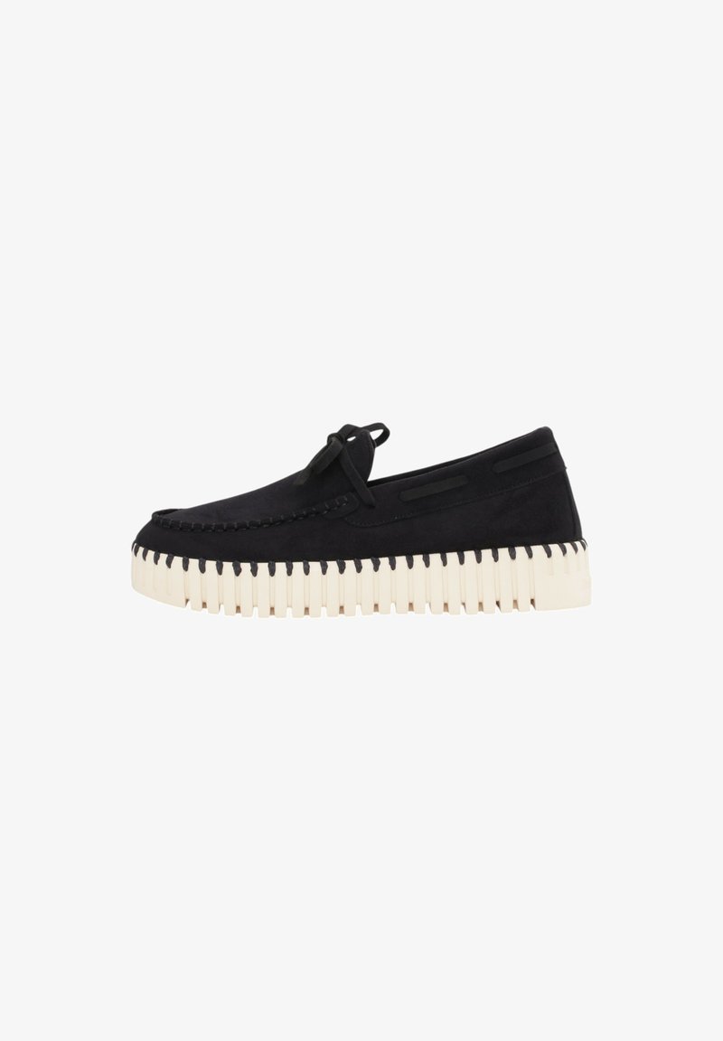Zapatos de ante negros sin cordones con una suela beige acanalada y costuras decorativas. Presentan un adorno de lazo en la parte superior.