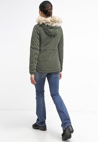 Oliwkowa kurtka z kapturem, wykończona sztucznym futrem, z ściąganym w pasie, zapinana na zamek, noszona na niebieskie dżinsy i czarne obuwie.